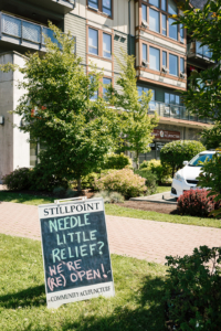 Sandwich board sign on a grass boulevard that reads "Needle Little Relief?"